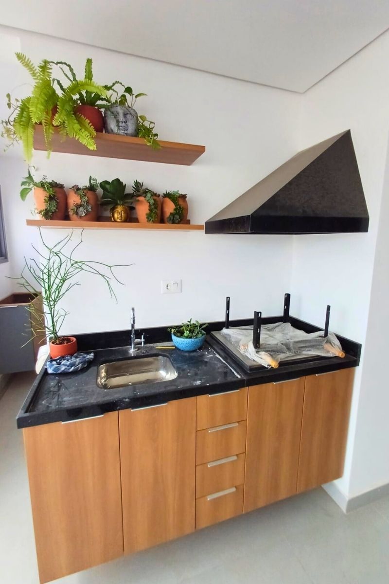 Cozinha moderna com bancada de granito preto, coifa de parede e prateleira superior decorativa em madeira com plantas.