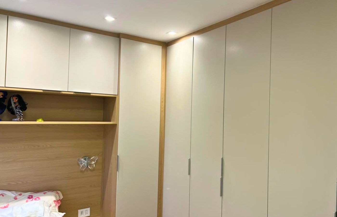 Quarto planejado de canto em tons claros (bege/branco), com armários em L otimizando o espaço do dormitório.