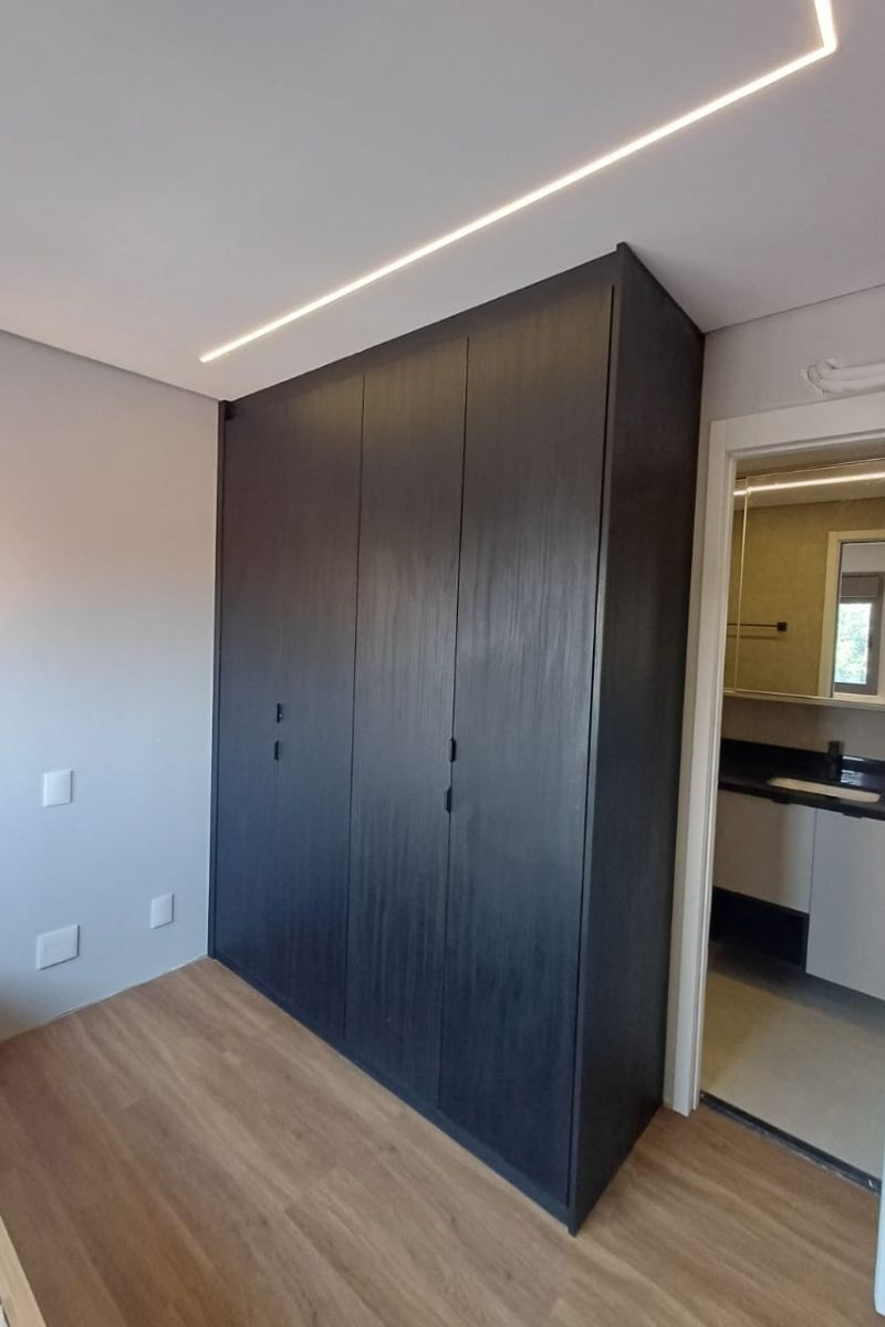 Roupeiro planejado em MDF preto (ou madeira escura texturizada) instalado em suíte, aproveitando o espaço ao lado da porta do banheiro.