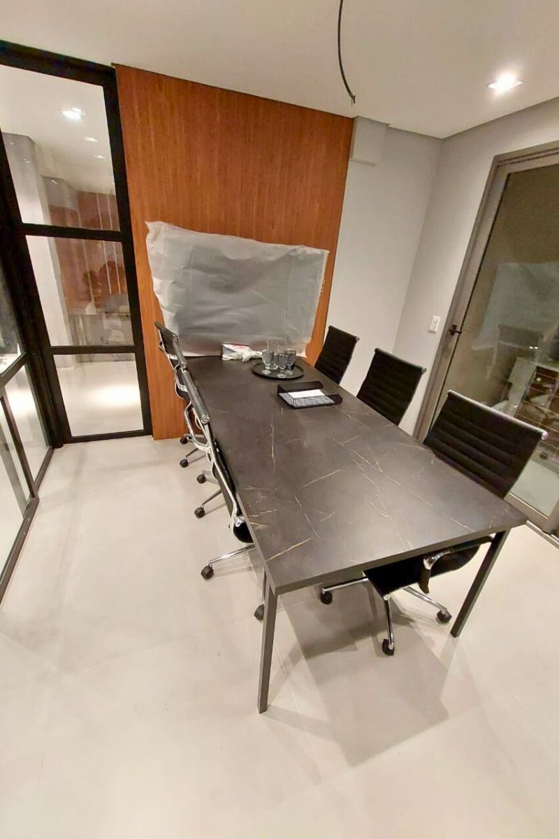 Mesa de reunião com tampo em padrão mármore escuro e cadeiras pretas em sala com divisória de vidro.