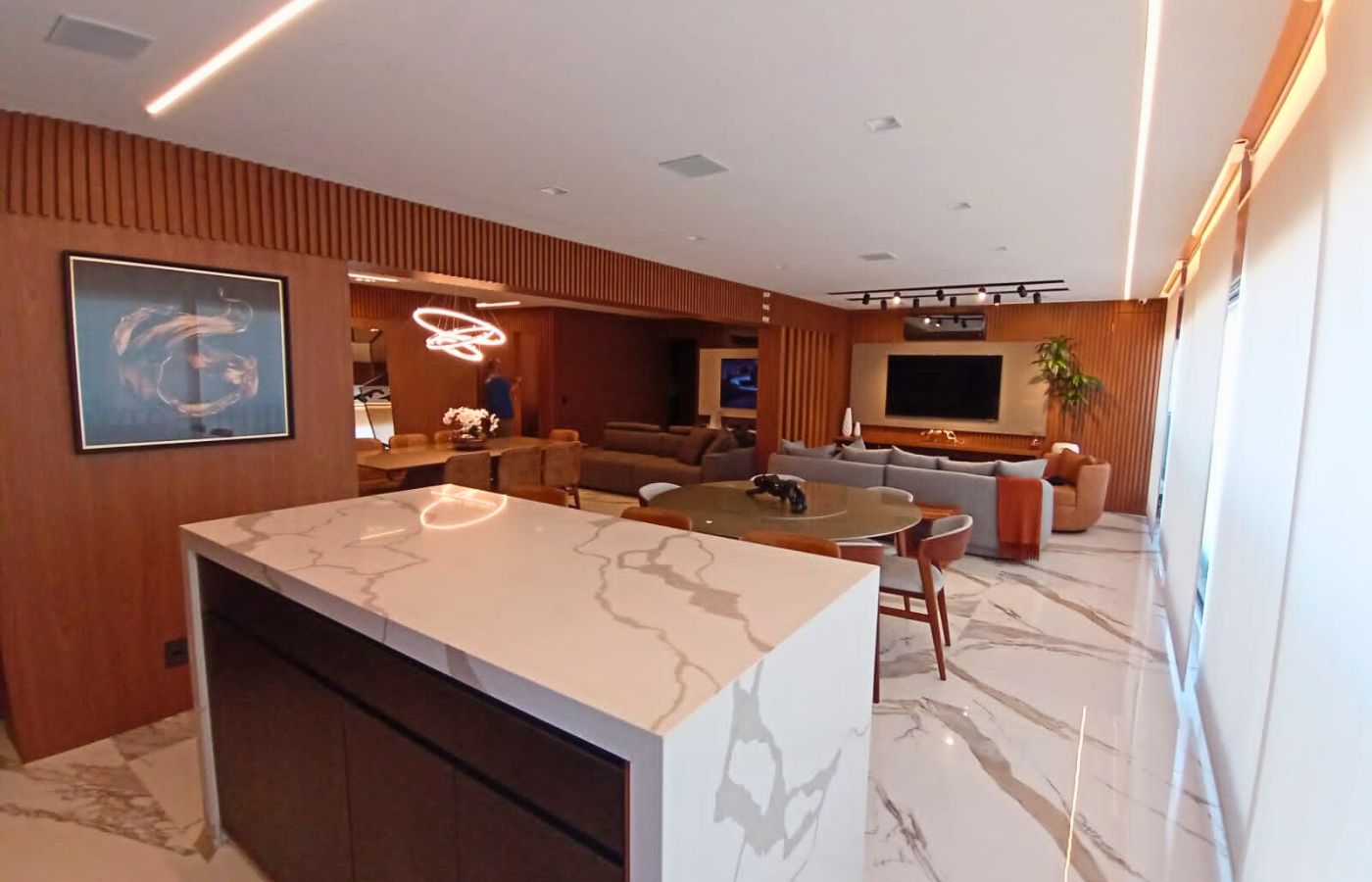 Ambiente integrado de alto padrão: cozinha com ilha de mármore branco conectada a uma sala de estar decorada com móveis planejados em madeira.
